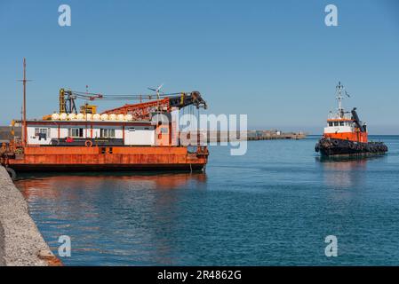 Candia, Creta, Grecia. 2023. Rimorchiatore che si avvicina ad una grande chiatta di gru a mare con una gru montata sul ponte a fianco del porto. Foto Stock