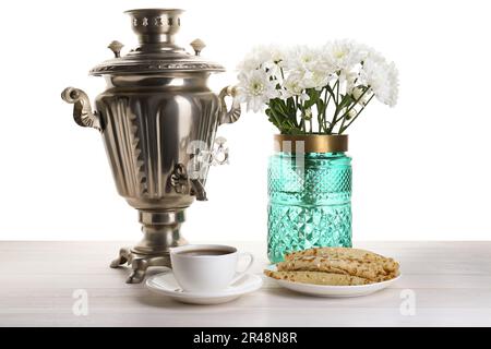 Samovar vintage, tazza di bevanda calda, frittelle e vaso con fiori su tavolo di legno su sfondo bianco. Tradizionale cerimonia del tè russo Foto Stock