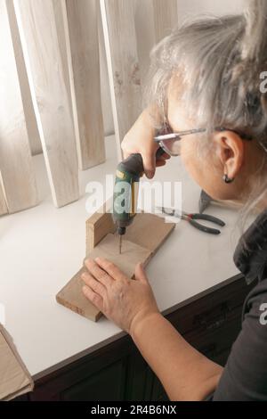 Donna vista in profilo con un cacciavite elettrico che mette le viti in un pezzo di legno Foto Stock