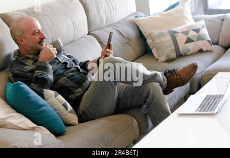 Uomo anziano con baffi e camicia a quadri seduto sul divano a casa bere caffè mentre guarda il suo telefono cellulare Foto Stock