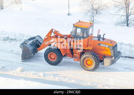 Dicembre 23, 2021. Regione di Kemerovo, Russia. Il trattore arancione grande pulisce la neve dalla strada e la carica nel carrello. Pulizia e pulizia della strada Foto Stock