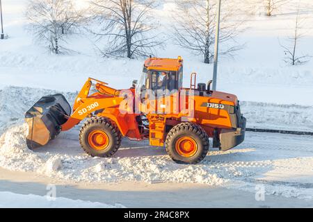 Dicembre 23, 2021. Regione di Kemerovo, Russia. Il trattore arancione grande pulisce la neve dalla strada e la carica nel carrello. Pulizia e pulizia della strada Foto Stock