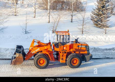 Dicembre 23, 2021. Regione di Kemerovo, Russia. Il trattore arancione grande pulisce la neve dalla strada e la carica nel carrello. Pulizia e pulizia della strada Foto Stock