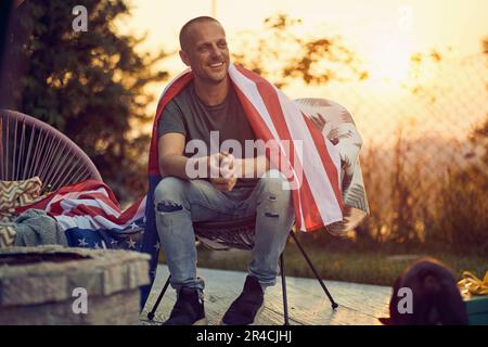 Ampio scatto orizzontale di giovane gioioso con la bandiera degli Stati Uniti sulle spalle, rilassandosi in una comoda sedia, all'aperto in natura. Concetto di Independence Day Foto Stock