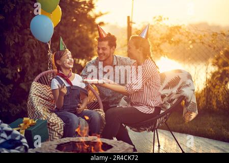 Il momento della famiglia mentre festeggiano il compleanno di loro figlia.la madre tiene una torta di compleanno bella, mentre il volto della figlia è giocosamente coperto dentro Foto Stock