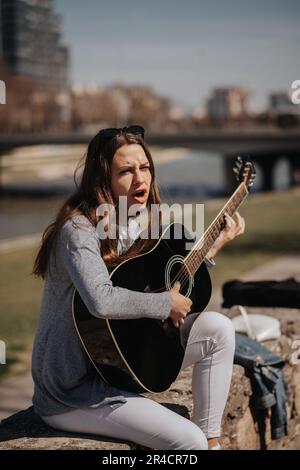 Bellissima ragazza brunetta seduta sulla banchina e suonando la chitarra. Sta cantando ad alta voce Foto Stock
