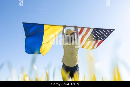 Donna in giorno di sole si alza con la schiena e solleva due grandi bandiere - americano e ucraino Foto Stock