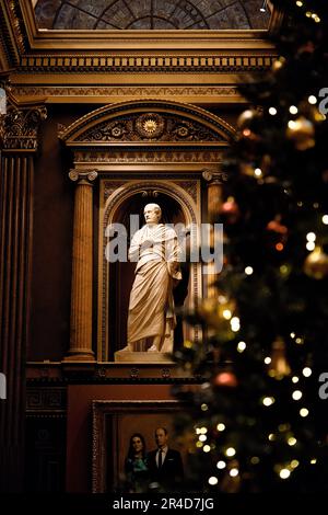 Un gruppo di persone si riunisce in una sala decorata, con un grande albero di Natale festivo nell'angolo, una statua decorativa e un grande soffitto a cupola Foto Stock