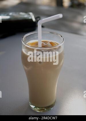 Uno sfondo sfocato e un caffè ghiacciato in un bicchiere su un tavolo di plastica con una cannuccia bianca. Foto Stock