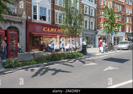Gente fuori e circa lo shopping in Marylebone High Street durante il giorno soleggiato di inizio estate. I commensali si siedono ai tavoli del marciapiede fuori dal ristorante italiano Carlotta. Foto Stock