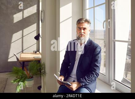 Uomo bearded grigio di mezza età con capelli seduto dalla finestra in ufficio Foto Stock