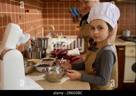 Bella bambina in cappello e grembiule dello chef, aiuta sua madre a fare una torta di festa mescolando panna montata con cioccolato fuso Foto Stock