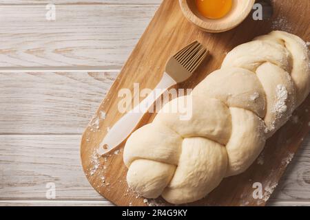 Pane crudo intrecciato, tuorlo d'uovo e pennello da pasticceria su tavolo di legno bianco, vista dall'alto con spazio per il testo. Tradizionale sfida Shabbat Foto Stock