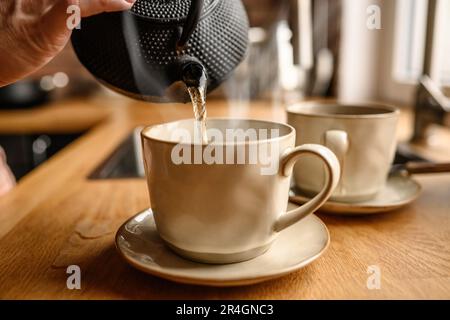 Persona che versa l'acqua calda dal bollitore nella tazza in cucina per preparare il tè. Teiera e tazza in ceramica con bevanda calda Foto Stock