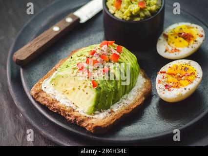 Toast integrale con avocado e formaggio cremoso e uova bollite. Serve con limone fresco e su piatto scuro. Vista dall'alto con primo piano. Foto Stock
