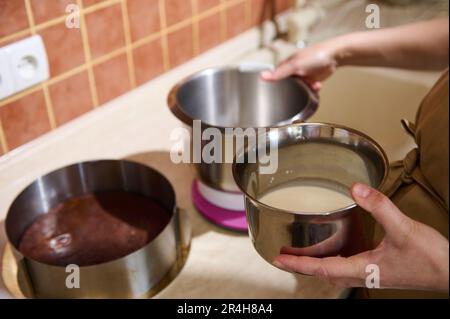 Primo piano delle mani della casalinga che tengono la ciotola con latte fresco o crema sopra un piano cucina. Cucina. Pasticceria Foto Stock