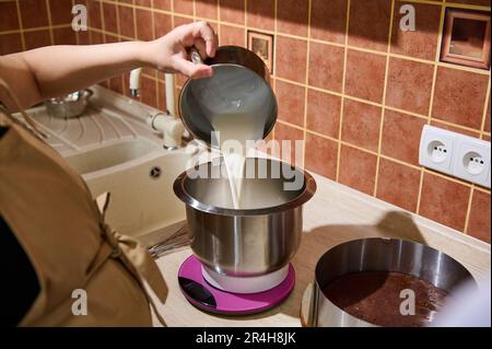 Mani ravvicinate di una casalinga, pasticciere femminile che versa panna fresca in una ciotola di robot da cucina con mixer planetario Foto Stock