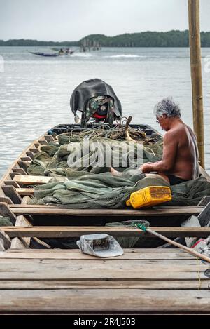 Chantaburi, Thailandia, 21 marzo 2023: Un pescatore in una barca di legno ripara la sua rete di pesca mentre una barca a coda lunga passa in lontananza. Foto Stock