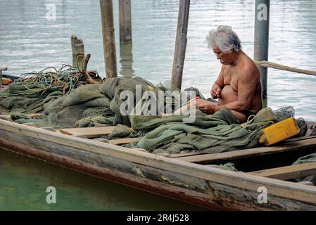 Chantaburi, Thailandia, 21 marzo 2023: Un pescatore siede all'interno della sua barca di legno che ripara una rete da pesca. Foto Stock