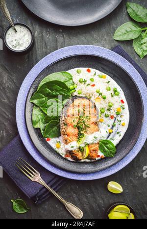 Piatto di bistecca di salmone alla griglia servito con spinaci per bambini, salsa di yogurt all'aglio, riso bianco, limone ed erbe fresche su sfondo scuro. Vista dall'alto con primo piano Foto Stock