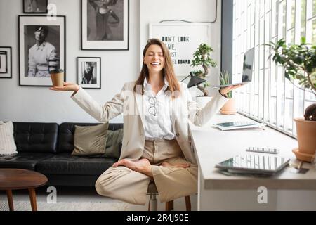Donna imprenditore felice meditare, seduta in yoga posa armi fuori diffusione, tenendo in mano laptop e caffè Foto Stock