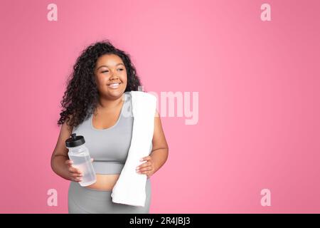 Donna allegra nera Plus size in abbigliamento sportivo con asciugamano che tiene una bottiglia d'acqua, riposante dopo l'allenamento su sfondo rosa Foto Stock