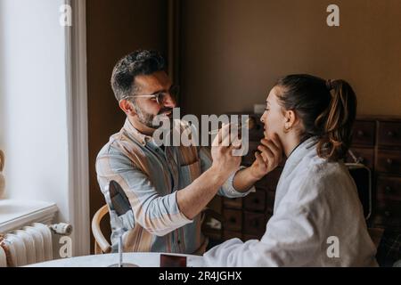 Ragazzo sorridente che applica il make-up a una persona non binaria a casa Foto Stock