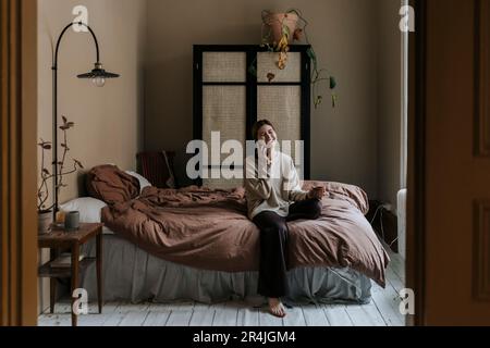 Felice non-binary persona che parla sullo smartphone mentre si siede in camera da letto a casa Foto Stock