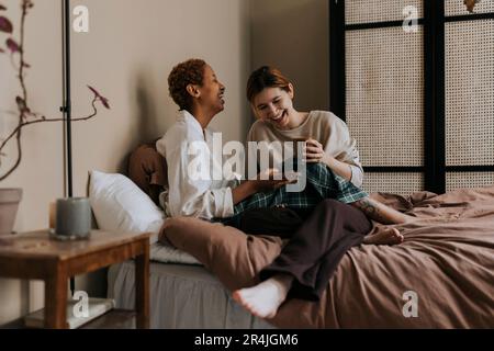 Felice coppia non binaria che condivide lo smartphone mentre si siede a letto a casa Foto Stock