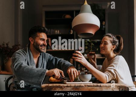 Felice non-binary persona che condivide smartphone con il ragazzo mentre fare colazione a casa Foto Stock