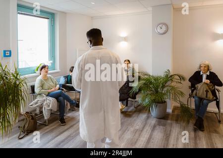Vista posteriore del medico maschile che parla con i pazienti seduti in sala d'attesa in ospedale Foto Stock