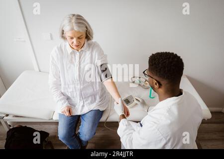 Vista ad angolo alto del medico di sesso maschile che misura la pressione sanguigna di un paziente anziano seduto a letto in clinica medica Foto Stock