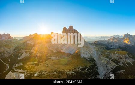 Il sole sorge da dietro la cima di una silhouette rocciosa di montagna. Paesaggio delle Alpi dolomitiche sotto il cielo limpido. Tre Cime di Lavaredo all'alba vista aerea sul retro illuminato Foto Stock