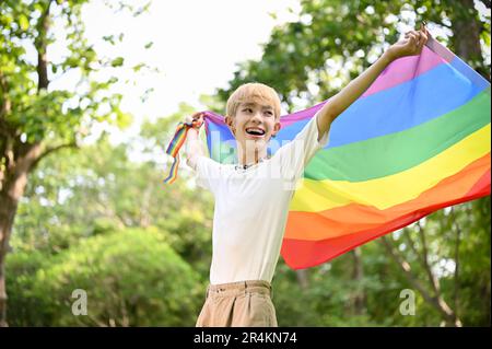 Ritratto di un giovane gay asiatico attraente e gioioso con una bandiera arcobaleno LGBT in piedi nel parco verde, sentendo libertà e orgoglio. Foto Stock