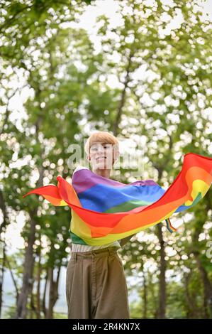 Ritratto di un giovane gay asiatico attraente e gioioso con una bandiera arcobaleno LGBT in piedi nel parco verde. Concetto LGBTQ+ Foto Stock