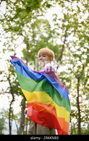 Ritratto di un giovane gay asiatico attraente e sorridente con una bandiera arcobaleno LGBT in piedi nel parco verde. Diversità, uguaglianza, diritti umani, Fre Foto Stock