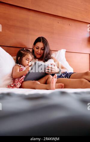 Una bambina e la sua giovane madre si divertono a usare un iPad sul letto della loro camera d'albergo Foto Stock