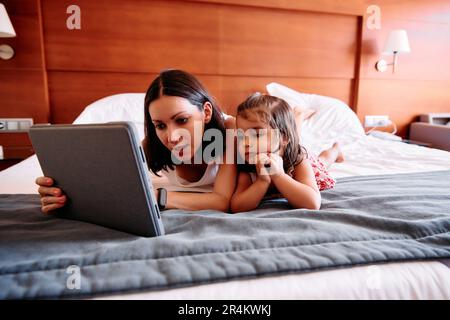 Una bambina e la sua giovane madre si divertono a usare un iPad sul letto della loro camera d'albergo Foto Stock