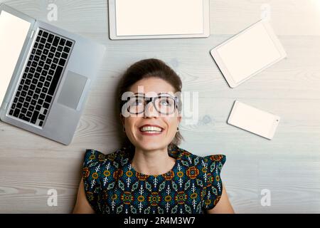 Donna felice in occhiali circondati da 4 schermi: Notebook, smartphone, tablet grandi e piccoli. Foto dall'alto che suggerisce la sua gioia con lo schermo cont Foto Stock