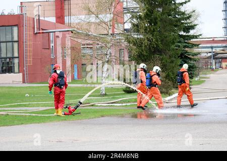 I vigili del fuoco professionisti in tute arancioni resistenti al fuoco in caschi bianchi con maschere a gas stanno testando manichette e pistole antincendio per spegnere un incendio Foto Stock