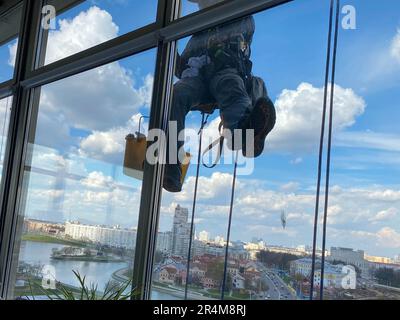 Un operatore di lavafinestre maschio, scalatore industriale pende da un alto edificio, grattacielo e lava grandi finestre di vetro per la pulizia alta sopra un lar Foto Stock