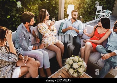 Sei giovani caucasici, tre uomini e tre donne, seduti in un elegante ambiente all'aperto, chiacchierando casualmente e gustando champagne. Lo scatto viene scattato fr Foto Stock