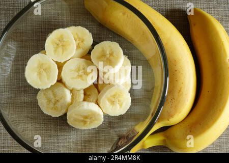 Fette di banana fresche mature in un recipiente di vetro e banane intere sul tavolo Foto Stock