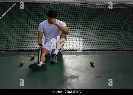 Giocatore asiatico di tennis maschile deluso seduto sul campo, turbato per la perdita di concorrenza, emozioni di fallimento Foto Stock