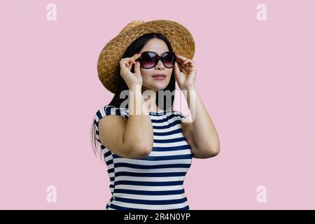 Giovane donna turistica asiatica indossa cappello di paglia e occhiali da sole. Isolato su sfondo rosa. Foto Stock