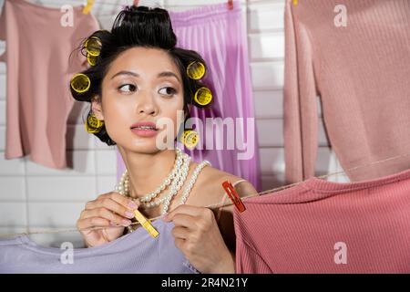 giovane donna asiatica pensiva con arricciacapelli e collana in perla che tiene spilla i vestiti e appeso lavanderia pulita e bagnata con sfondo sfocato a casa, Foto Stock