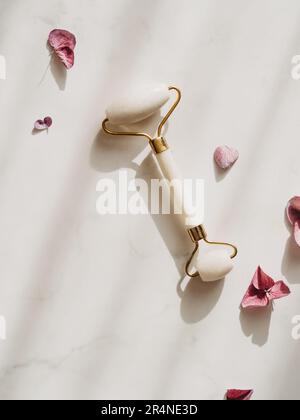 White Jade Face Roller per trattamenti di bellezza con massaggi facciali e fiori rosa di Hydrangea su sfondo bianco con ombre. Vista dall'alto. Spazio di copia. Foto Stock