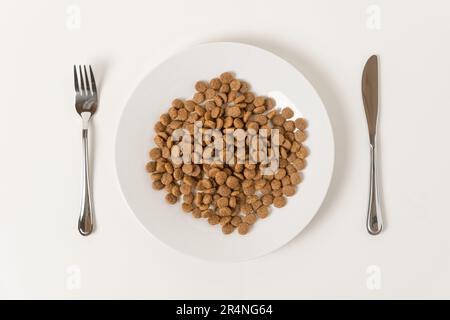 Cibo secco per animali in un piatto di ceramica bianca servito con coltello e forchetta su uno sfondo marrone neutro. Cibo per cani o gatti. Immagine divertente creativa per l'annuncio Foto Stock