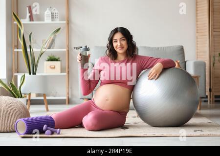Donna sportiva incinta con bottiglia d'acqua e fit ball a casa Foto Stock