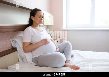 Donna in attesa negli ultimi stadi della gravidanza mettere le mani palme sul suo addome incinta, riposando a casa. Maternità Foto Stock